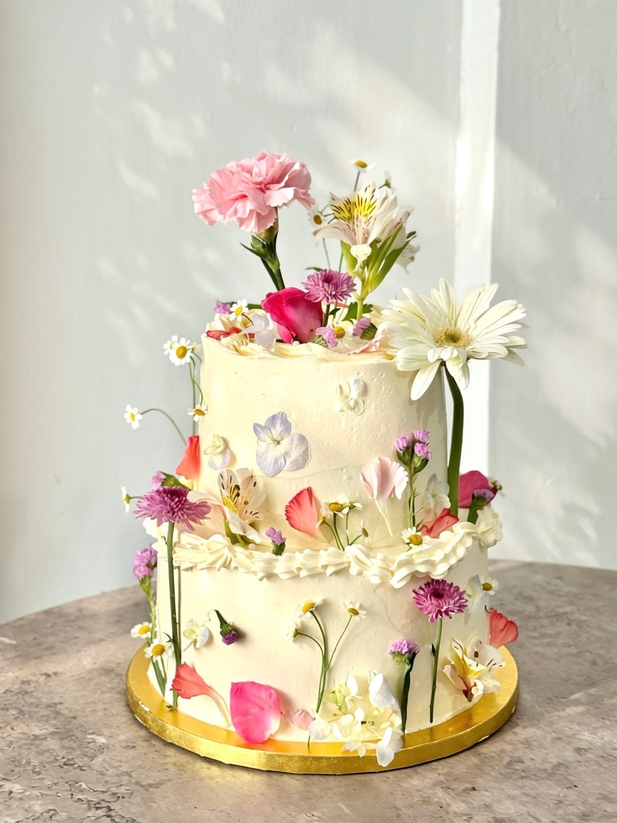 Two-tier white cake dressed with fresh carnations, daisies and petals